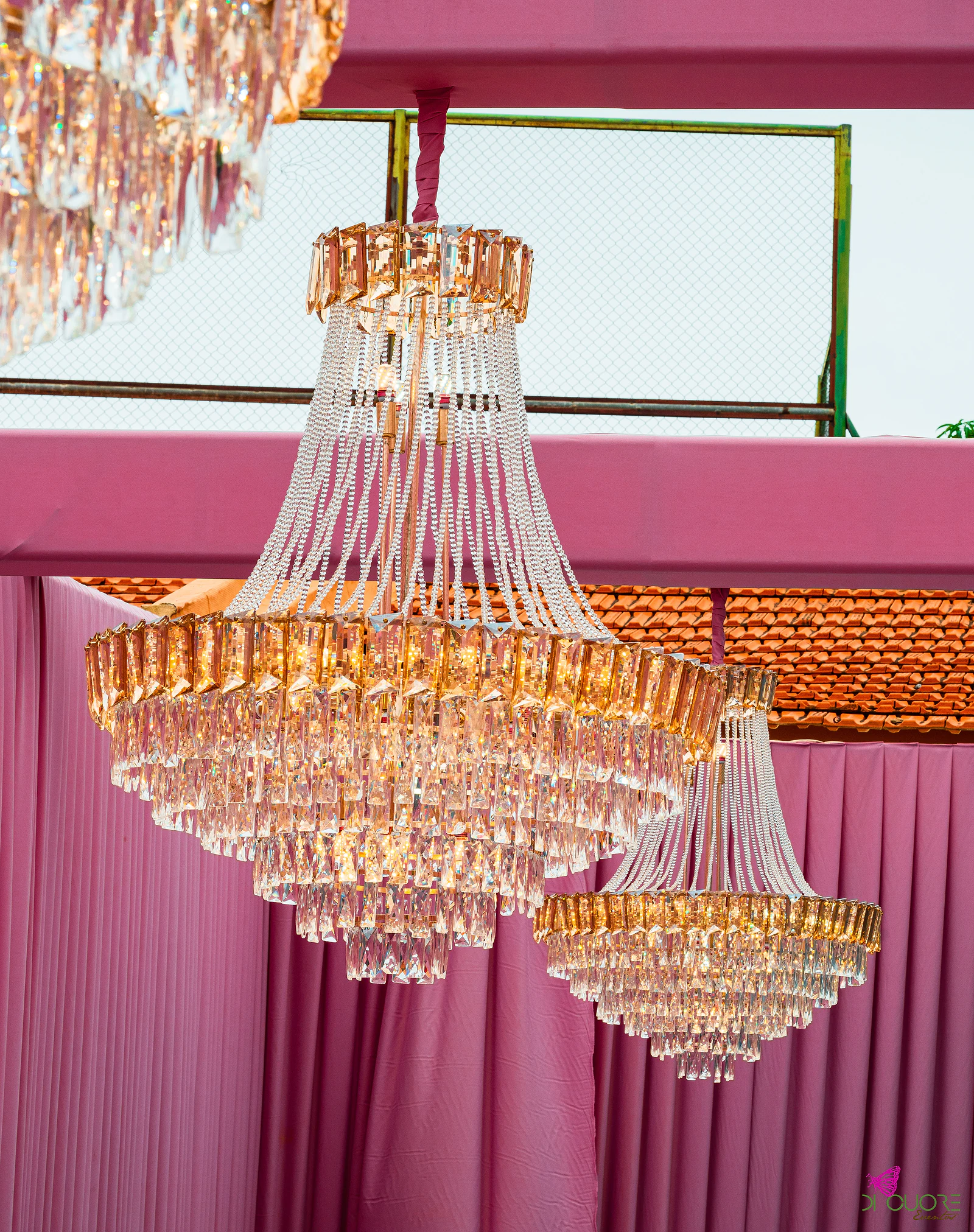 Dois lustres de cristal com pendentes dourados e fios de contas, suspensos diante de cortinados cor-de-rosa