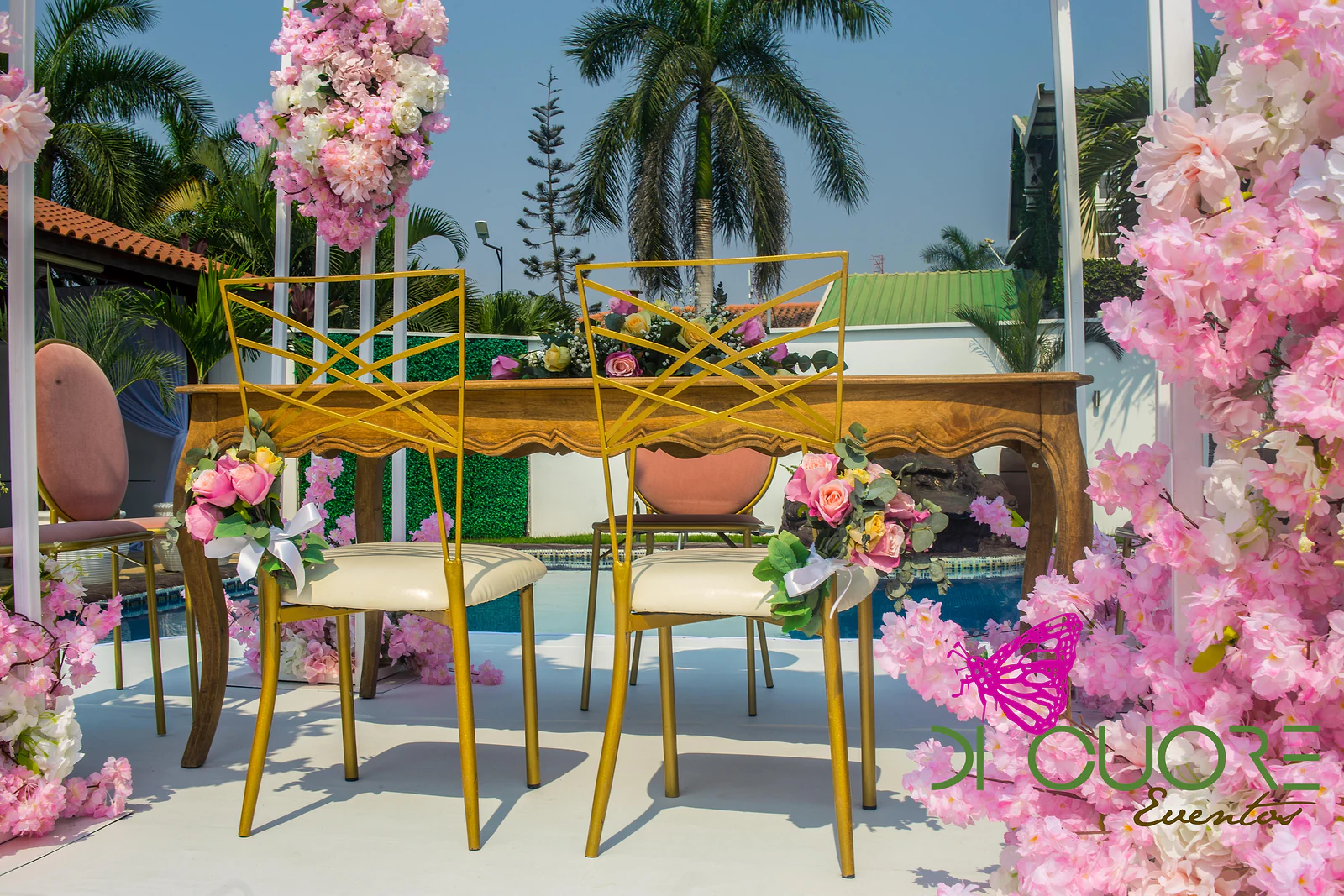 Mesa de madeira com duas cadeiras douradas enfeitadas com pequenos ramos de rosas cor-de-rosa e laços brancos, entre colunas de flores.