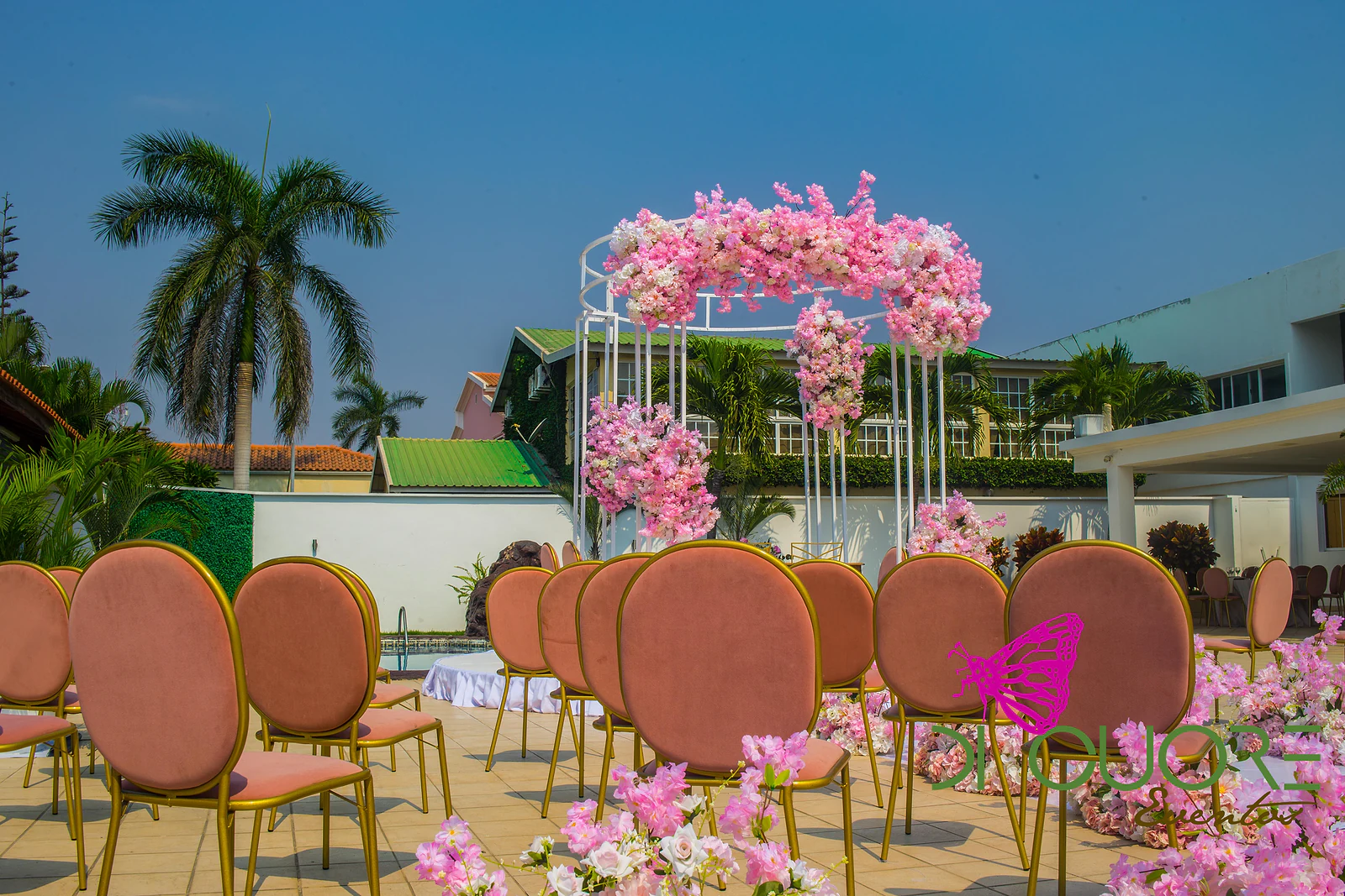Cadeiras de veludo cor de salmão com estrutura dourada em primeiro plano e, ao fundo, o pórtico de flores cor-de-rosa.