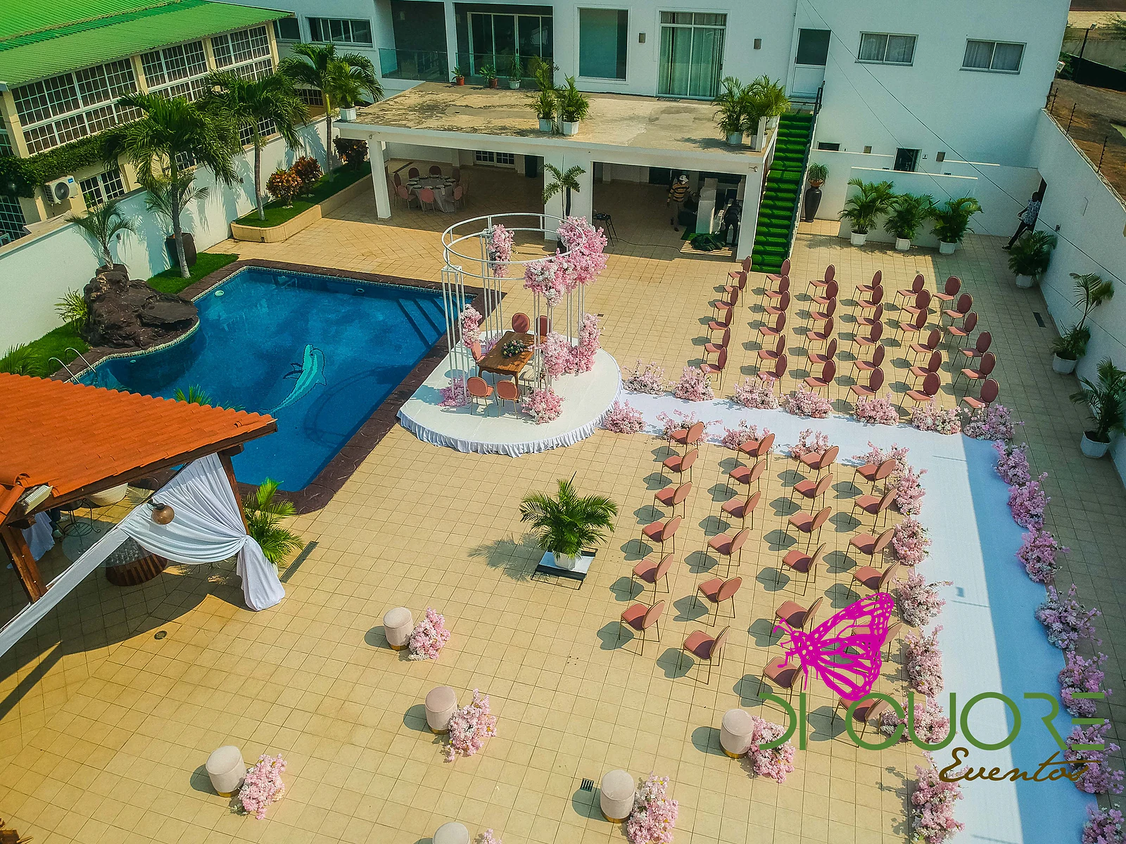 Vista aérea da cerimónia: fileiras de cadeiras cor de salmão em redor de uma passadeira branca, com o pórtico de flores e a piscina ao lado.
