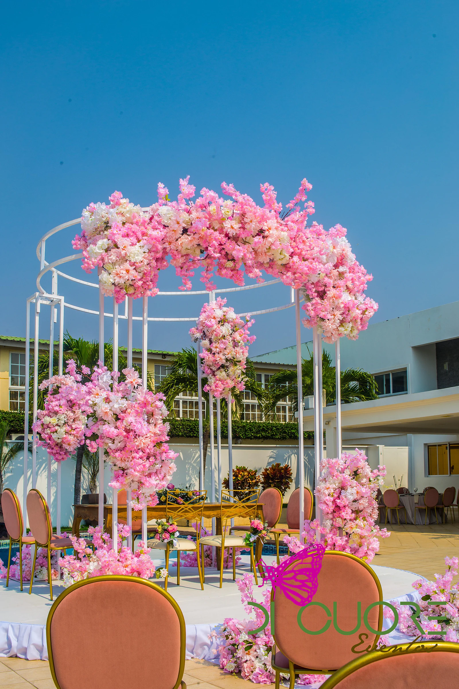 Estrutura branca coroada de flores cor-de-rosa e brancas, com a mesa de madeira ao fundo e cadeiras cor de salmão em primeiro plano.