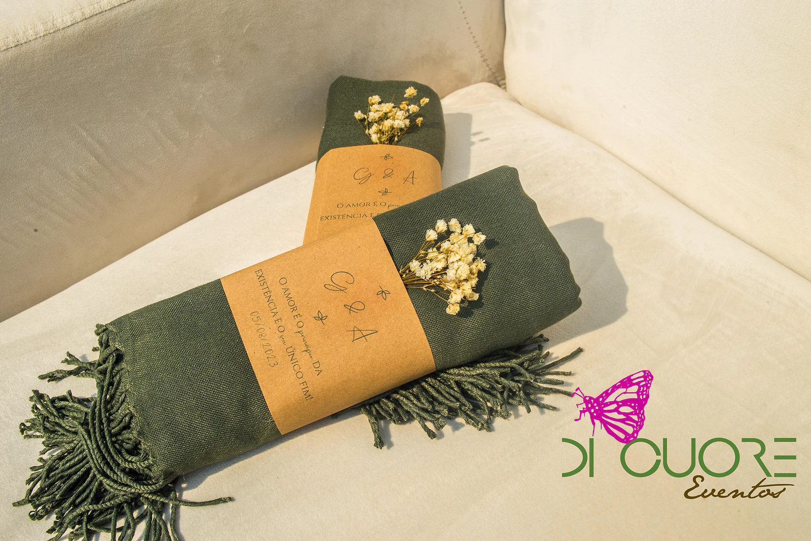 Dois xailes verde-escuro enrolados, cada um com uma cinta de papel craft com monograma e um raminho de gipsófila.