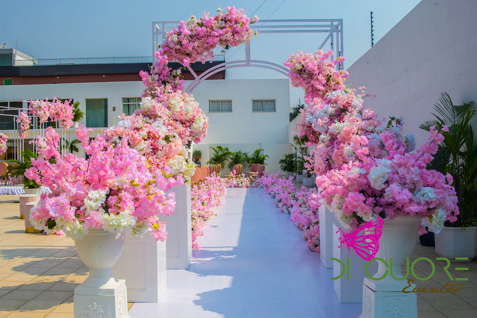 White aisle lined with white urns holding large pink and white floral arrangements, beside a floral arch on an open-air terrace