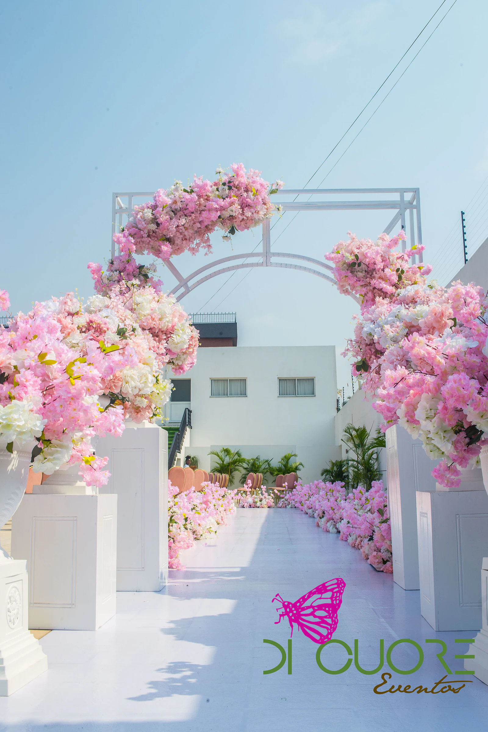 Passadeira branca entre urnas de flores cor-de-rosa e brancas sobre pedestais brancos, com um arco de flores ao fundo.