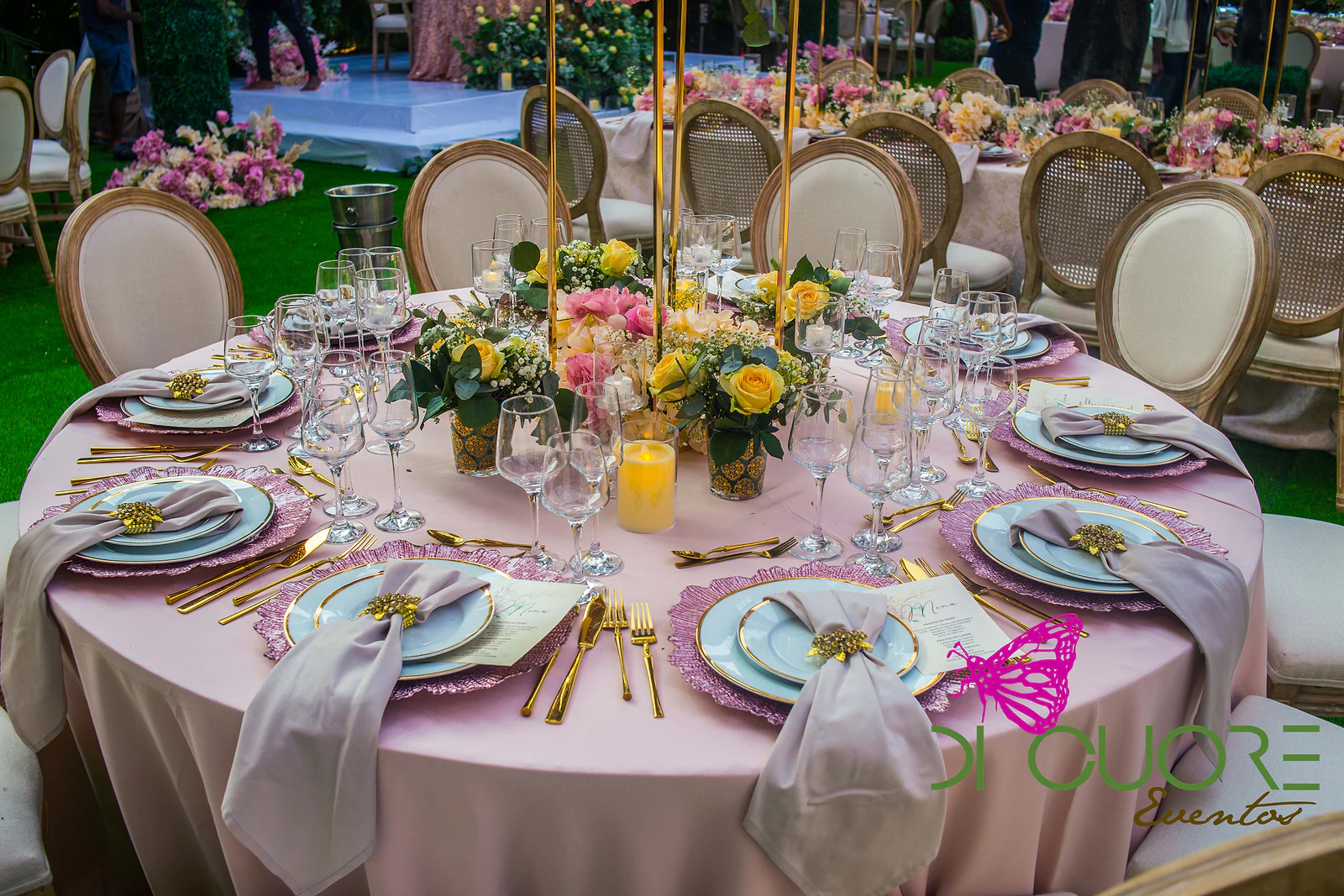 Mesa redonda com toalha cor-de-rosa, pratos azul-claro, guardanapos cor de malva e centro de mesa com rosas amarelas e velas.