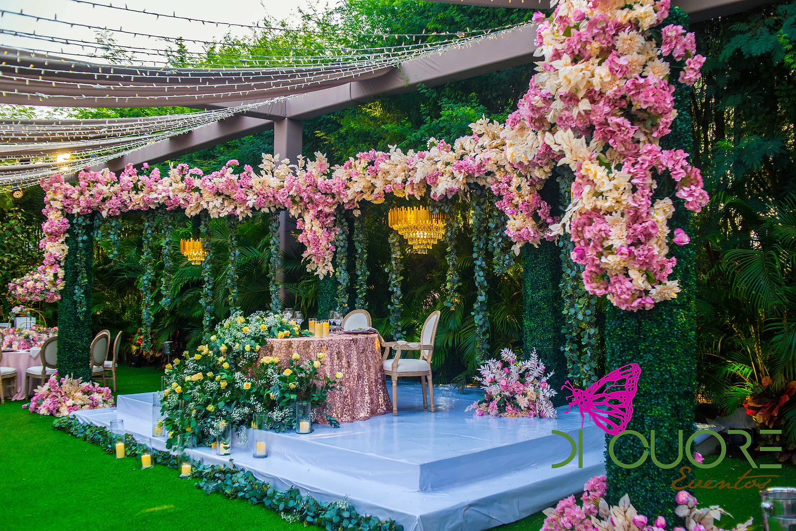 Estrado branco com mesa de lantejoulas cor-de-rosa e rosas amarelas, sob um pórtico de flores cor-de-rosa com lustres dourados, no jardim.