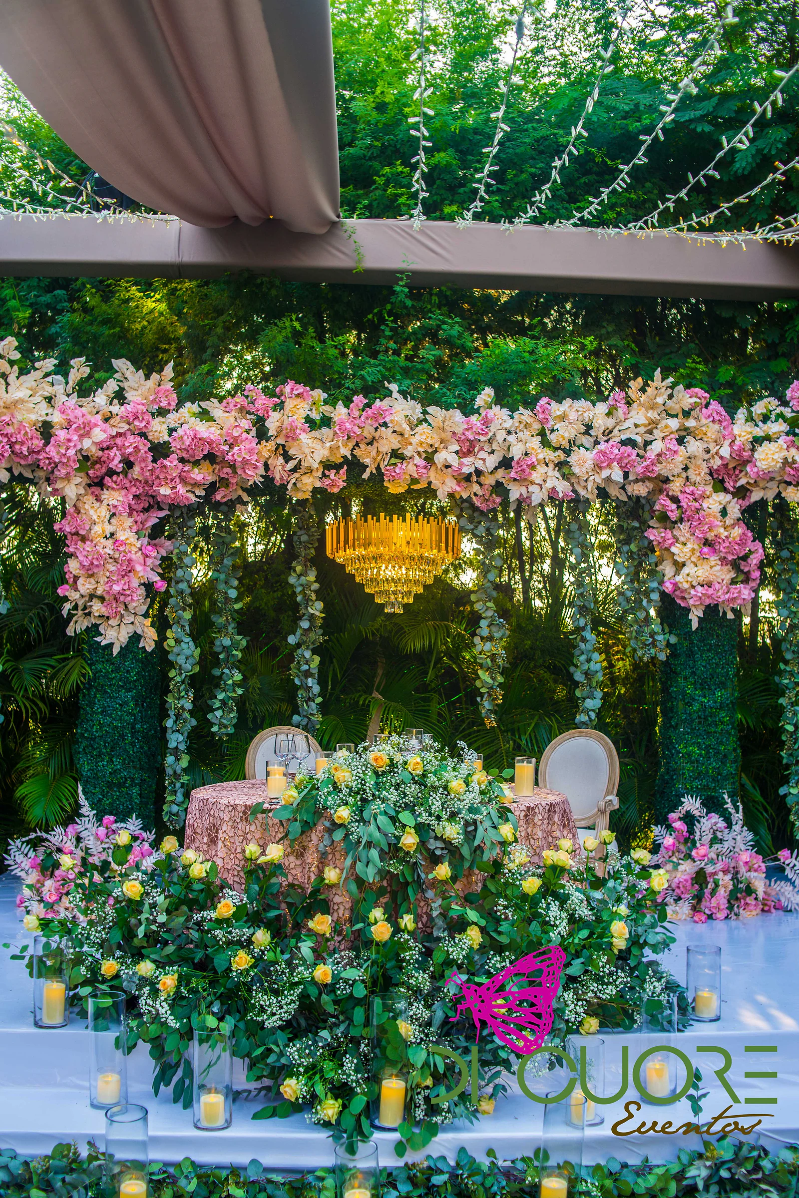 Mesa principal num estrado branco, rodeada de rosas amarelas, gipsófila e velas, sob um lustre dourado e uma cortina de flores cor-de-rosa.