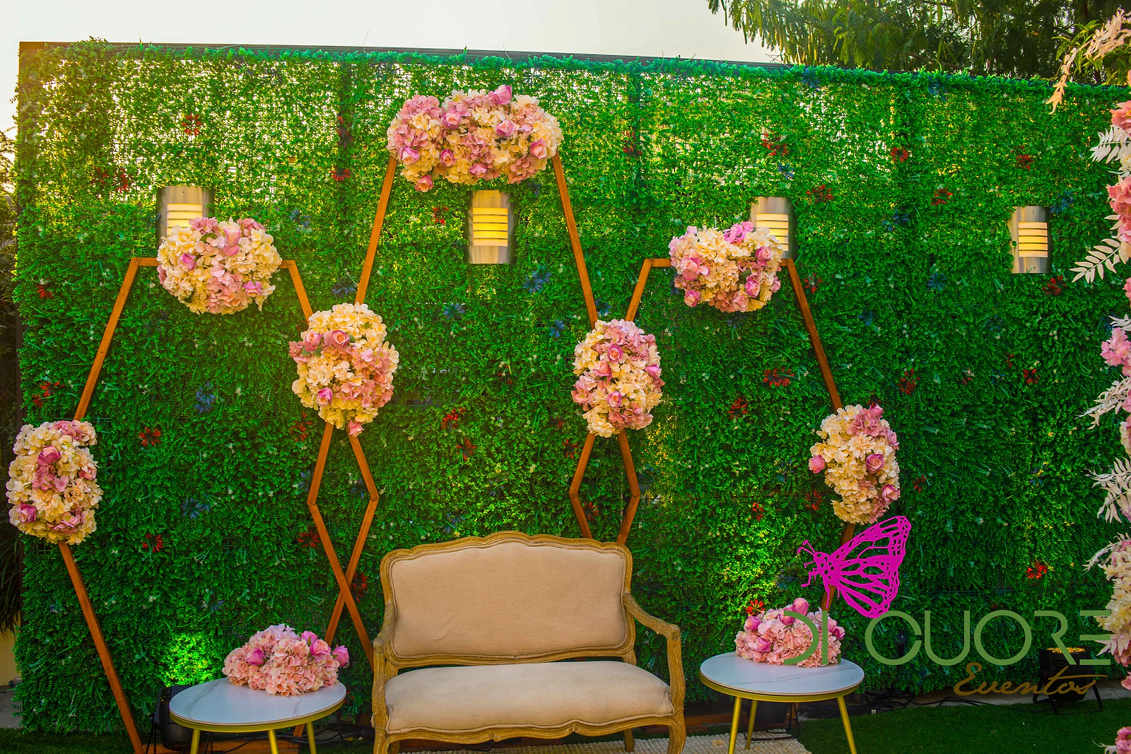 Painel de folhagem com estruturas hexagonais douradas e bolas de flores cor-de-rosa, com um sofá creme e duas mesas de apoio.