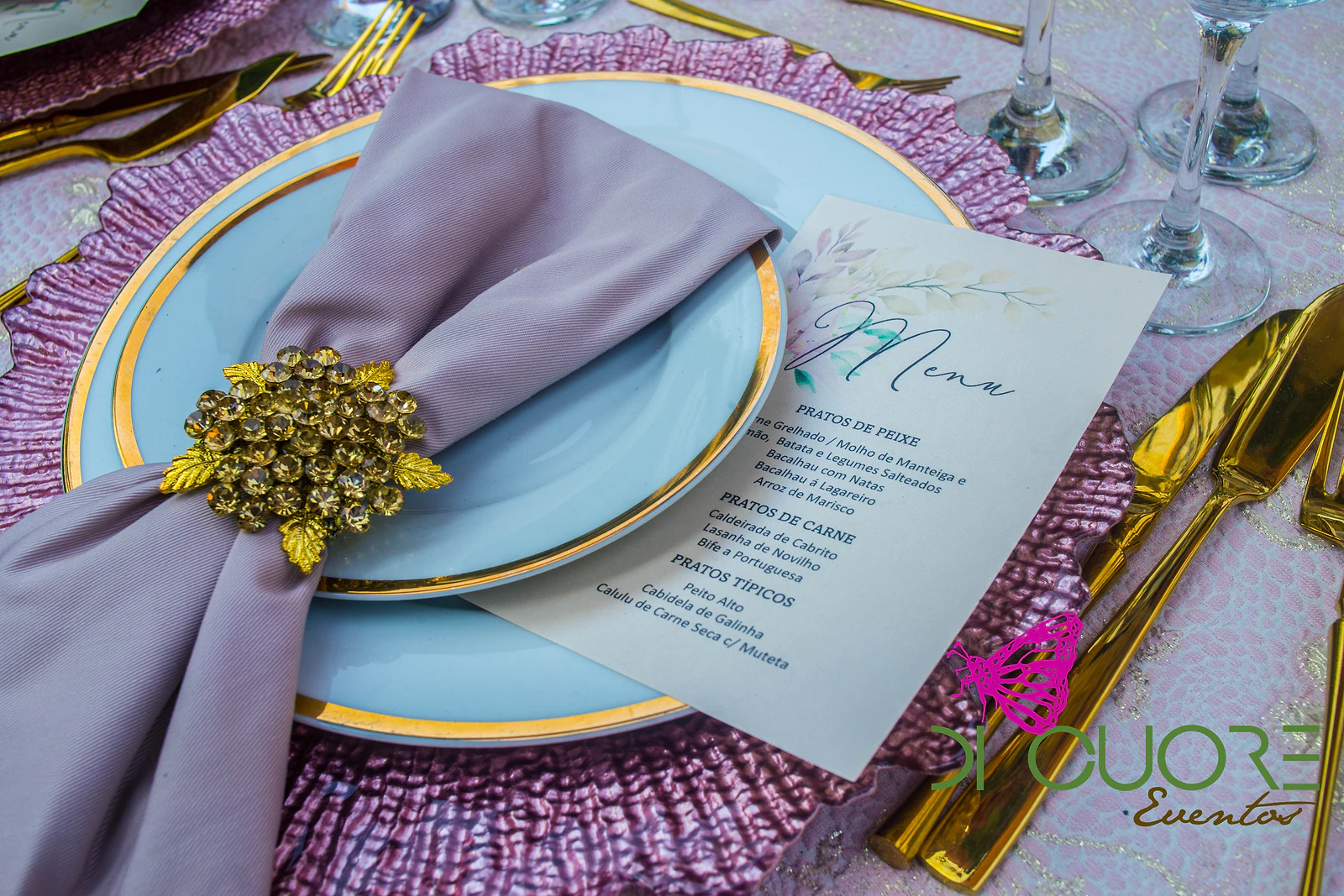Prato azul-claro com rebordo dourado sobre base cor-de-rosa, guardanapo cor de malva numa argola dourada e cartão de ementa.