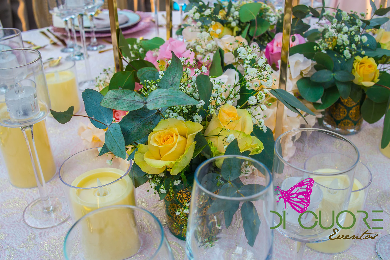 Rosas amarelas com gipsófila e folhas de eucalipto numa jarra, entre velas em copos de vidro.