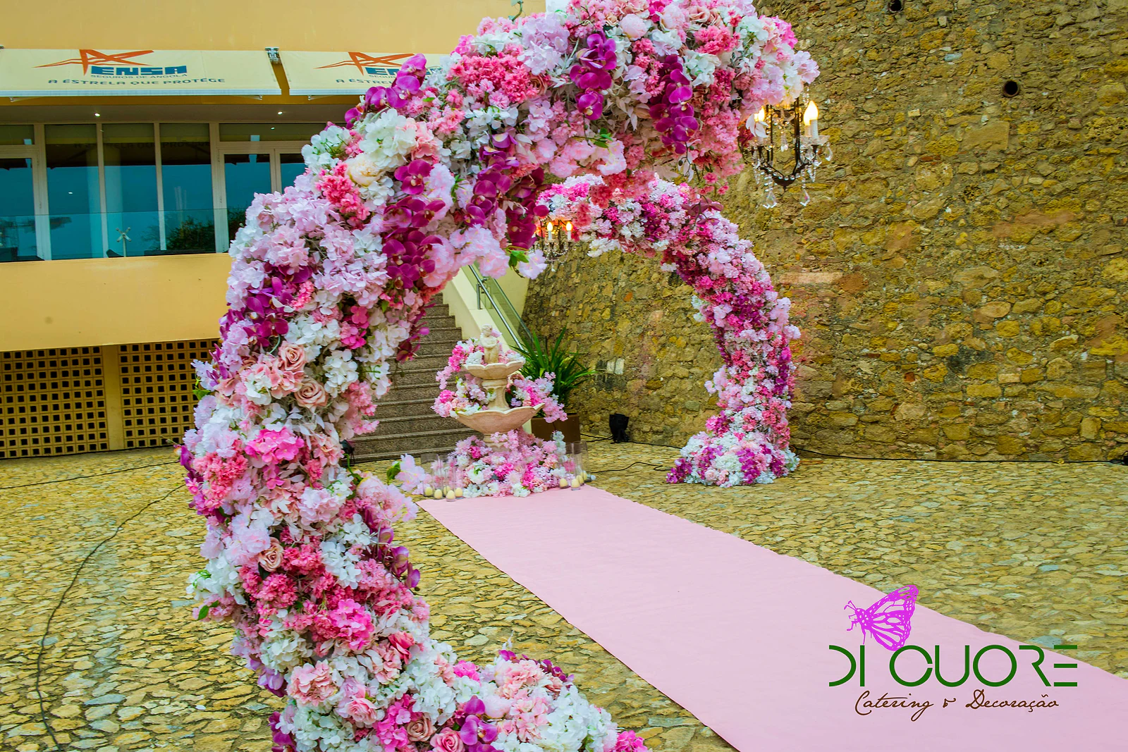 Arco curvo coberto de flores rosa, brancas e fúcsia sobre o tapete rosa, com uma fonte florida ao fundo junto a uma escadaria de pedra