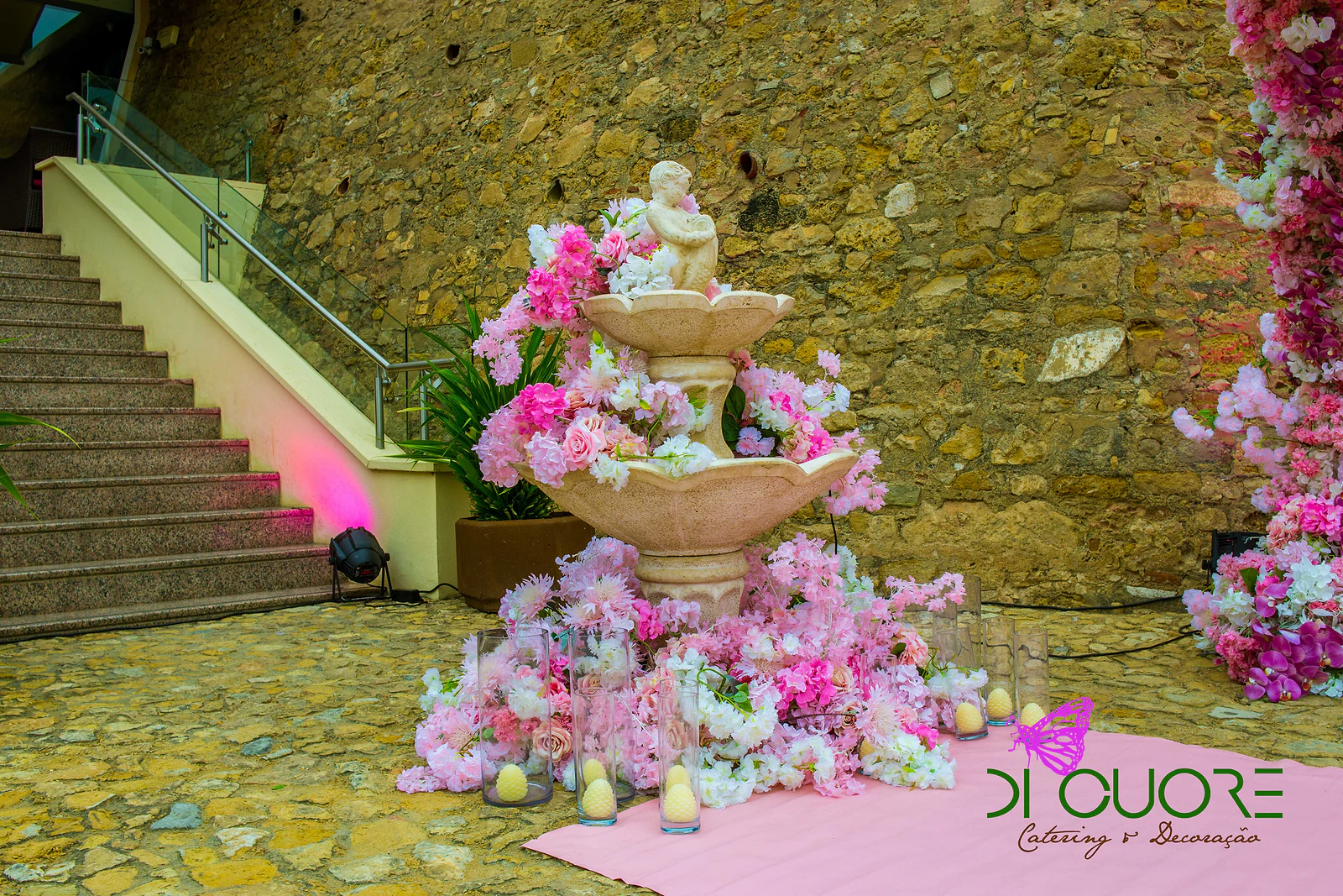 Fonte de dois pratos com uma figura de pedra, rodeada de flores cor-de-rosa e brancas e de velas em jarras de vidro, junto a uma escadaria de pedra