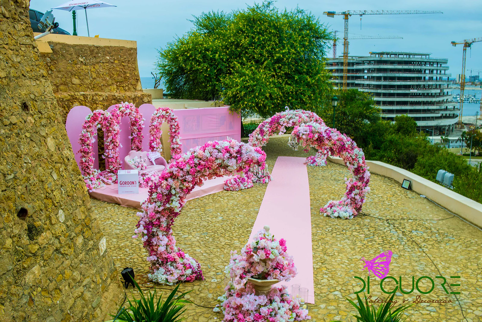 Tapete rosa pelo pátio de pedra entre arcos curvos de flores, com o estrado de arcos ao fundo e a cidade ao longe