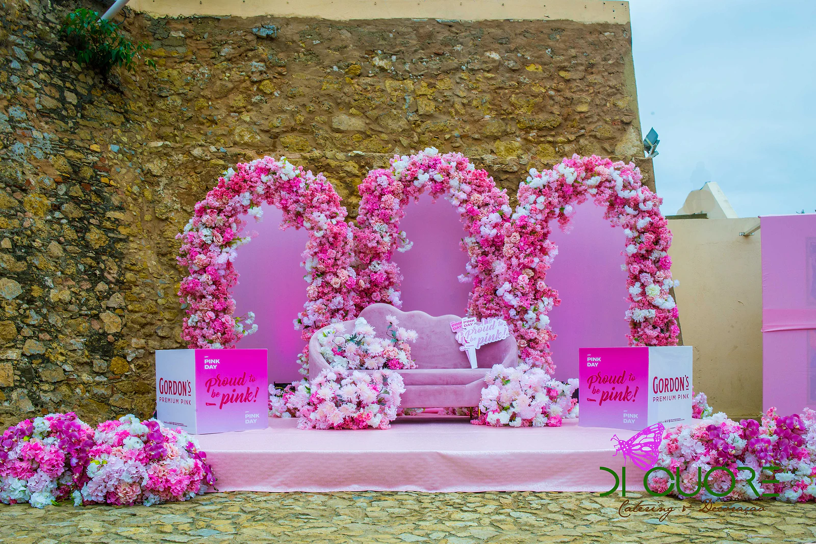 Três arcos de flores cor-de-rosa e brancas em frente a uma muralha de pedra, com um sofá rosa sobre estrado e cubos Gordon’s Premium Pink de cada lado