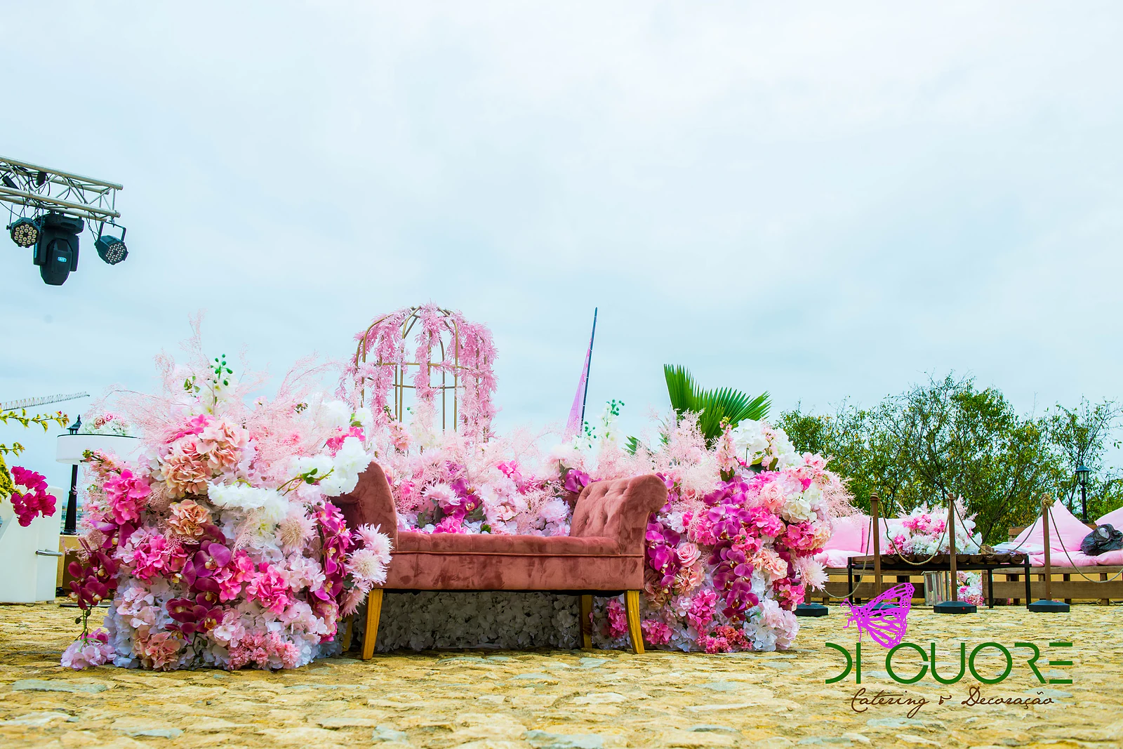 Banco de veludo rosado entre maciços de flores e ramos de plumas cor-de-rosa, com uma gaiola dourada coberta de flores ao fundo e o mar atrás