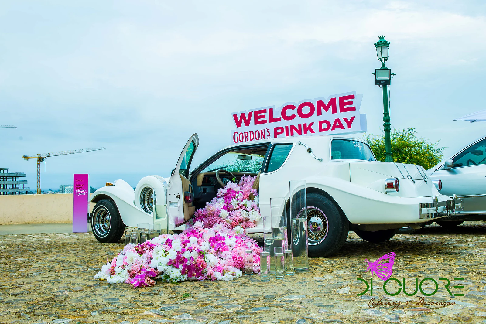 Carro antigo branco de portas abertas com o letreiro “Welcome Gordon’s Pink Day” e uma cascata de flores a escorrer para a calçada, junto a um candeeiro