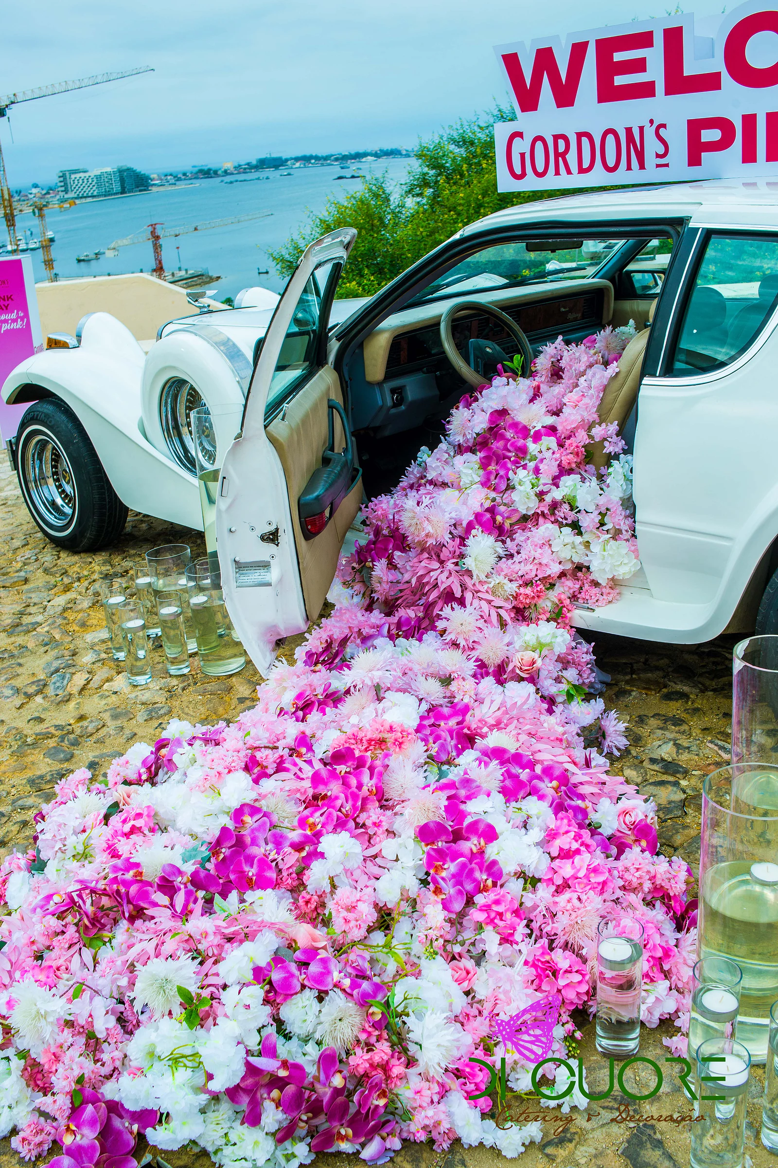 Cascata de orquídeas e flores fúcsia, rosa e brancas a escorrer de um carro antigo branco de portas abertas, com o letreiro de boas-vindas ao alto e a baía ao fundo