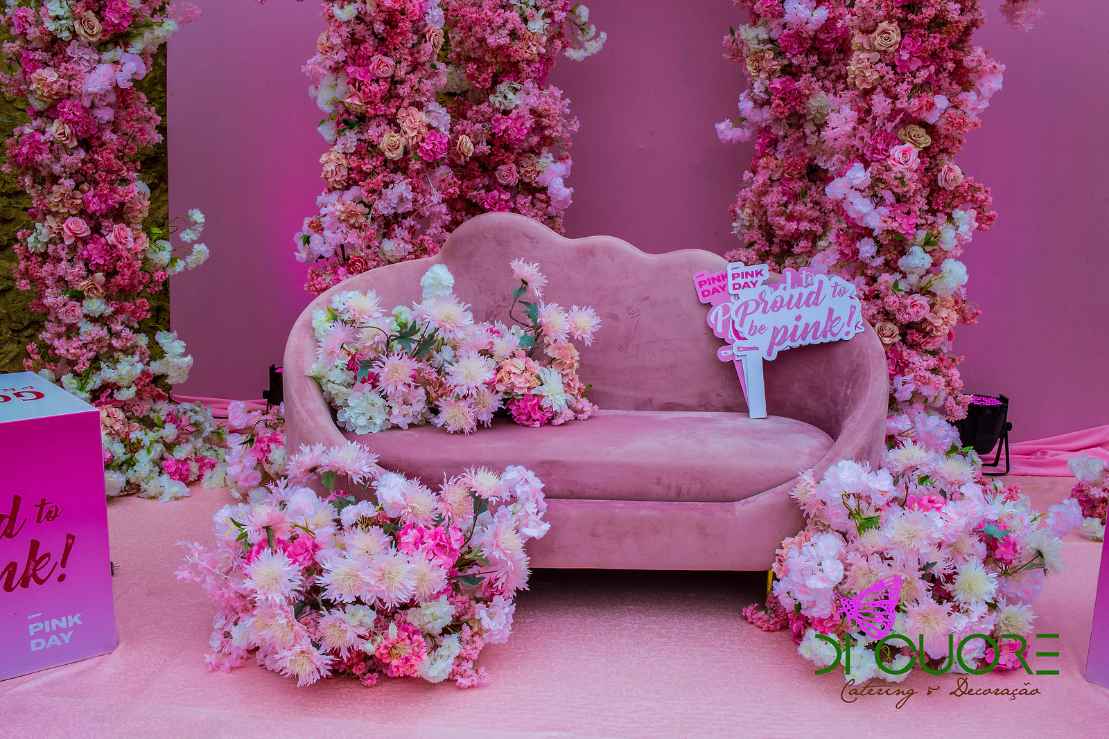Sofá de veludo rosa com ramos de flores nas almofadas e um cartaz “proud to be pink!”, diante de colunas de flores cor-de-rosa
