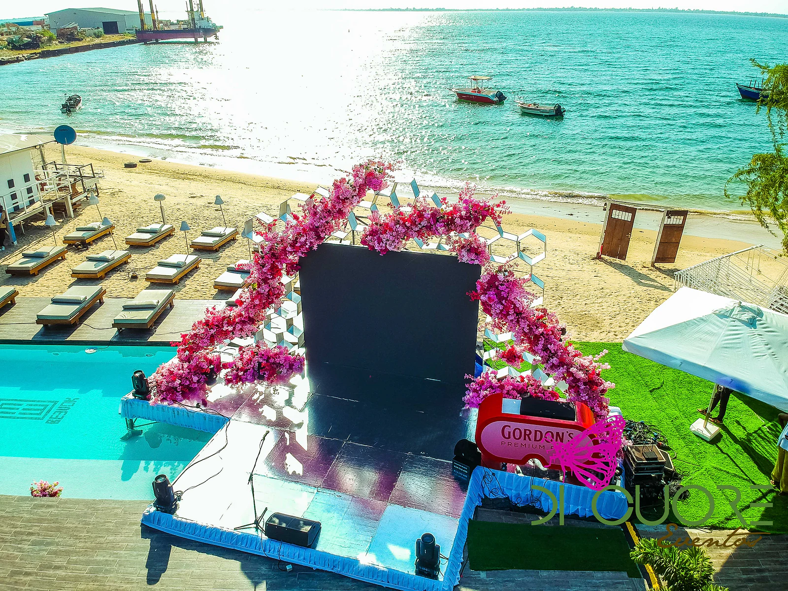 Vista aérea de um palco com um arco de flores cor-de-rosa e fúcsia, junto a uma piscina e a uma praia de areia com espreguiçadeiras, com o mar ao fundo