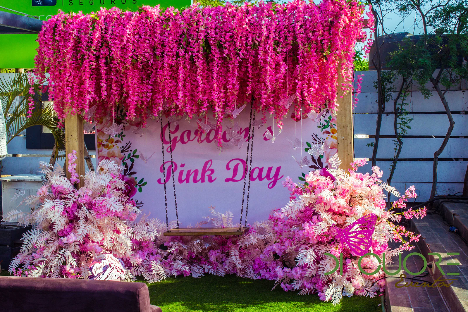 Cortina de flores fúcsia suspensas sobre um painel com o nome do evento e um baloiço, rodeado de folhagem branca e flores cor-de-rosa
