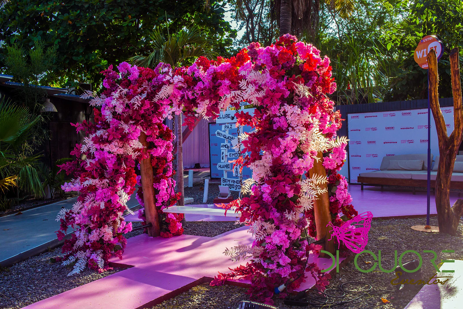 Arco de flores fúcsia, cor-de-rosa e brancas sobre um caminho rosa, entre árvores