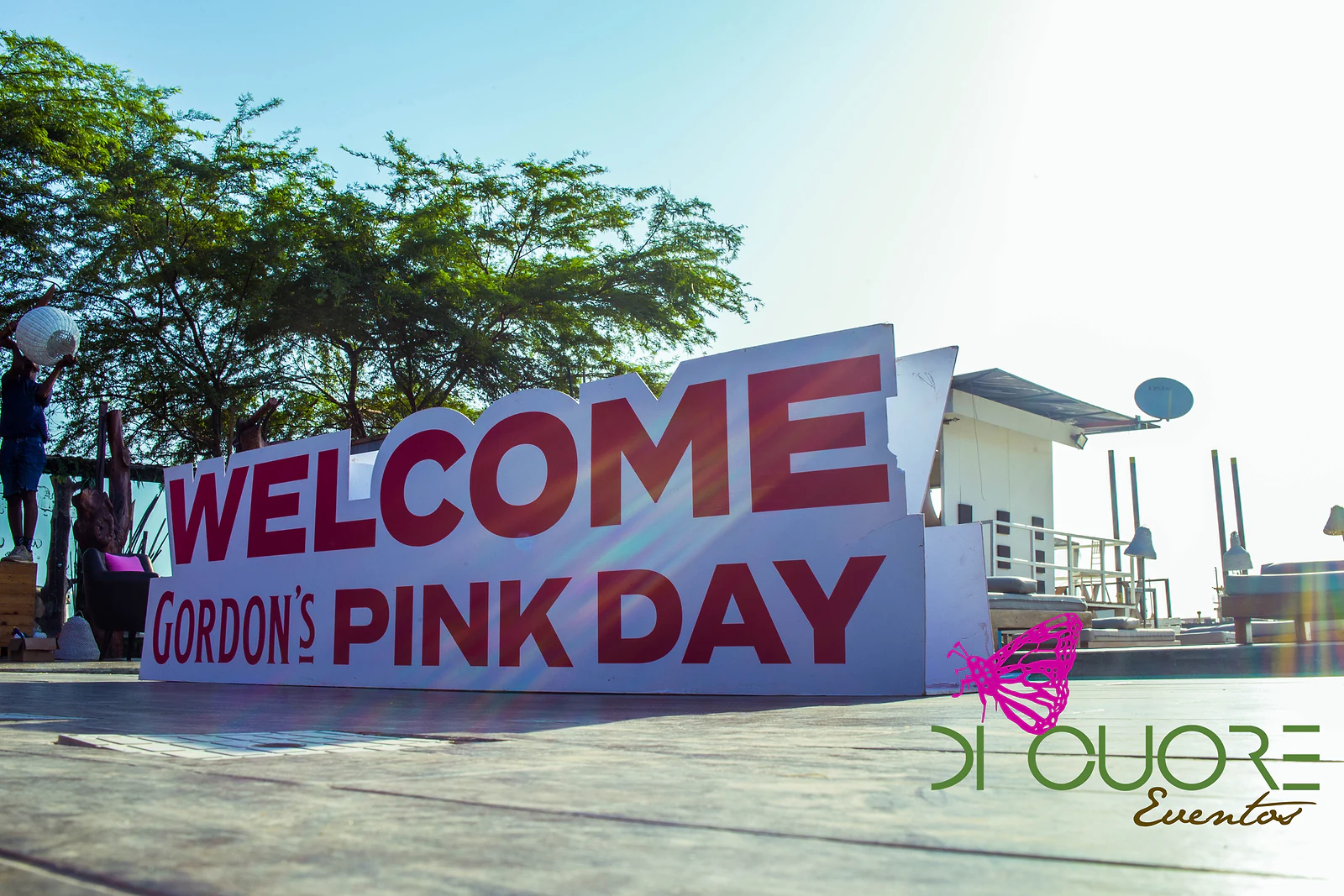 Letreiro de boas-vindas ao Gordon’s Pink Day, em letras vermelhas sobre fundo branco, junto ao deque e às árvores