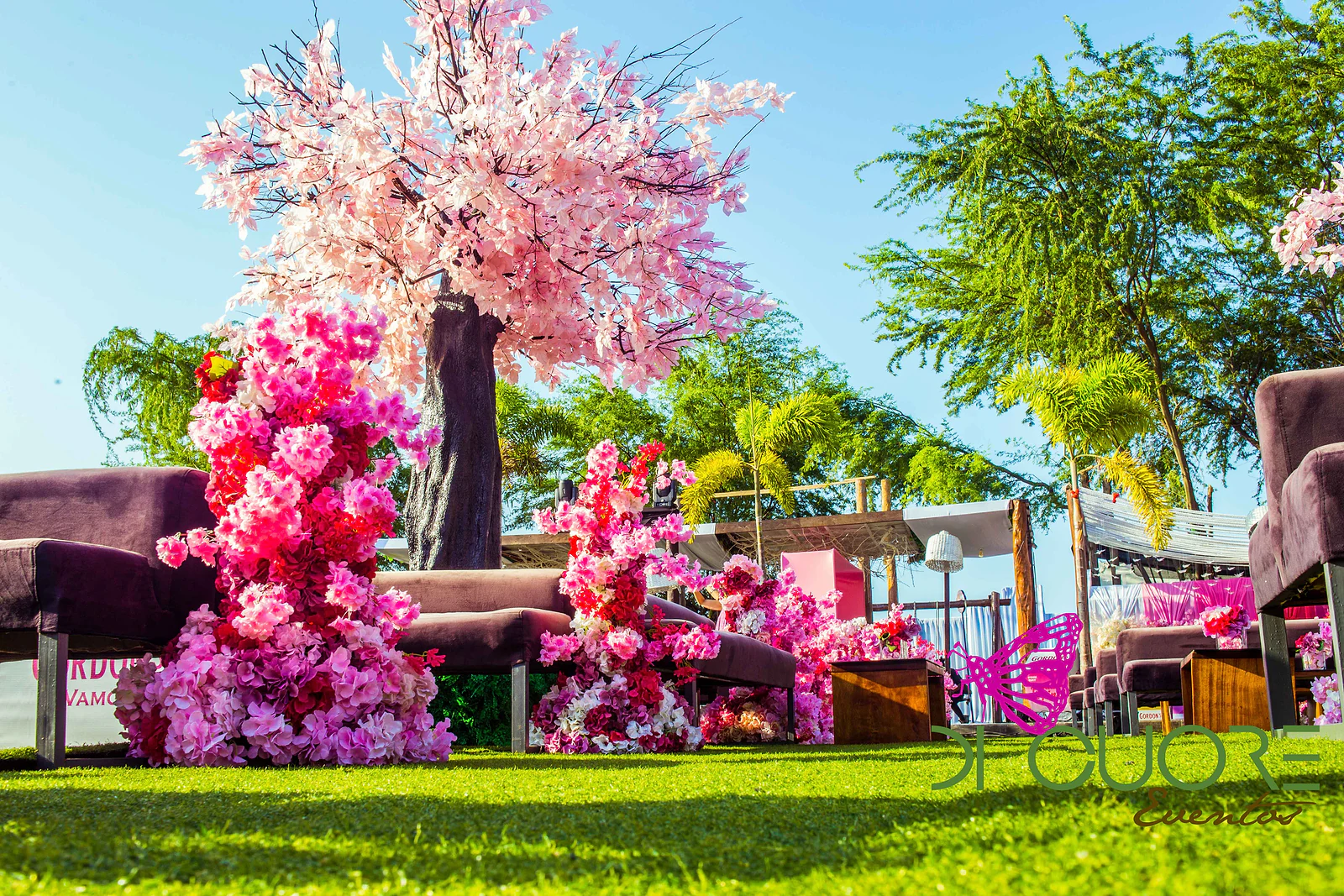 Cerejeira de flores cor-de-rosa pálido atrás de um maciço alto de flores fúcsia e cor-de-rosa, entre bancos de veludo, sobre o relvado