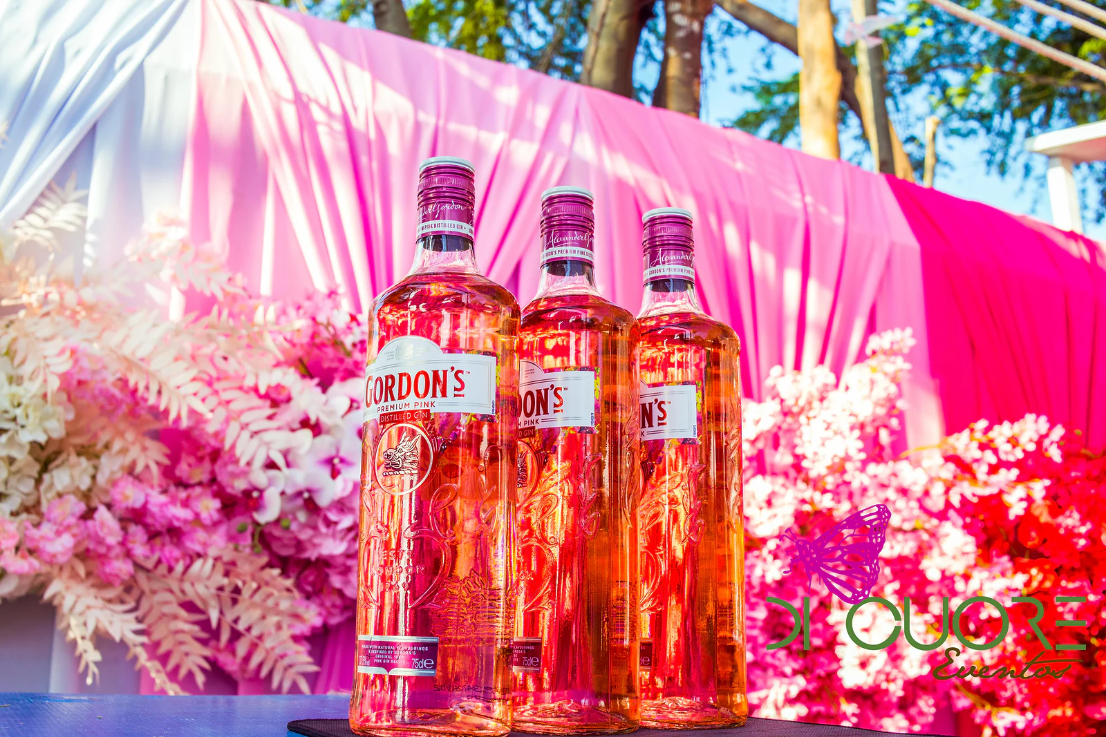 Três garrafas de Gordon’s Pink diante de panos cor-de-rosa e flores brancas, cor-de-rosa e vermelhas