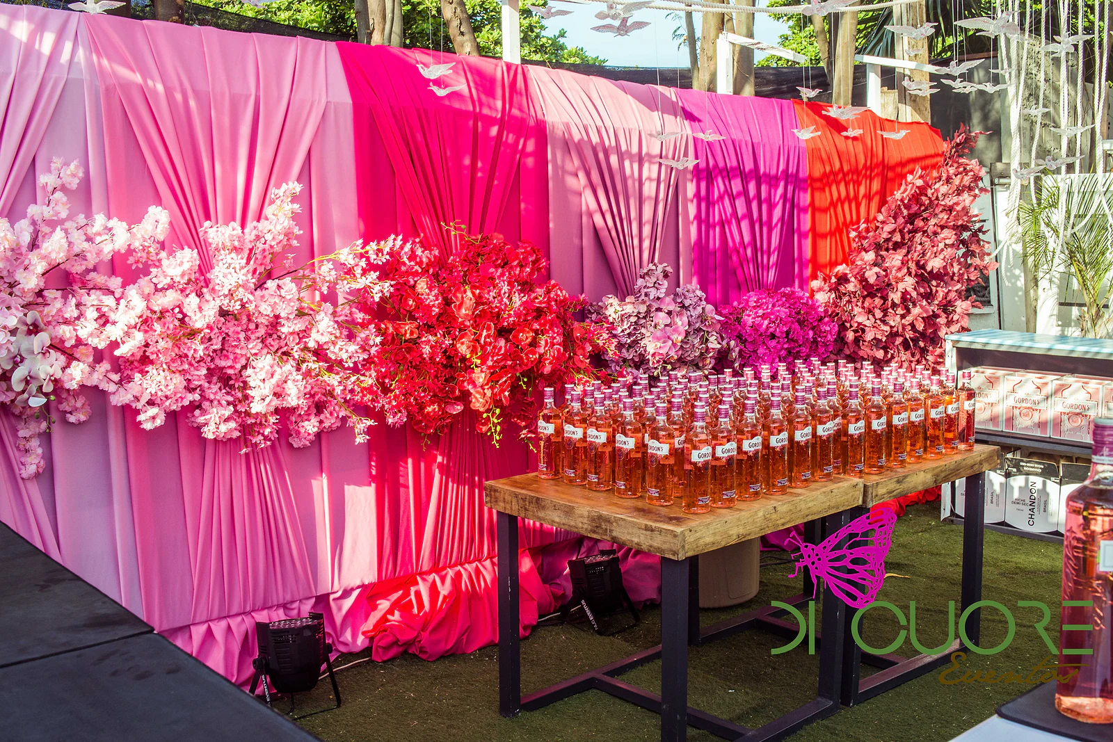 Balcão com panos drapeados do rosa-pálido ao vermelho, flores cor-de-rosa e fileiras de pequenas garrafas sobre uma mesa de madeira