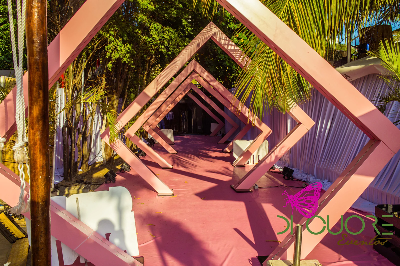 Corredor de losangos cor-de-rosa em madeira, com panos brancos e chão rosa, a perder-se entre palmeiras