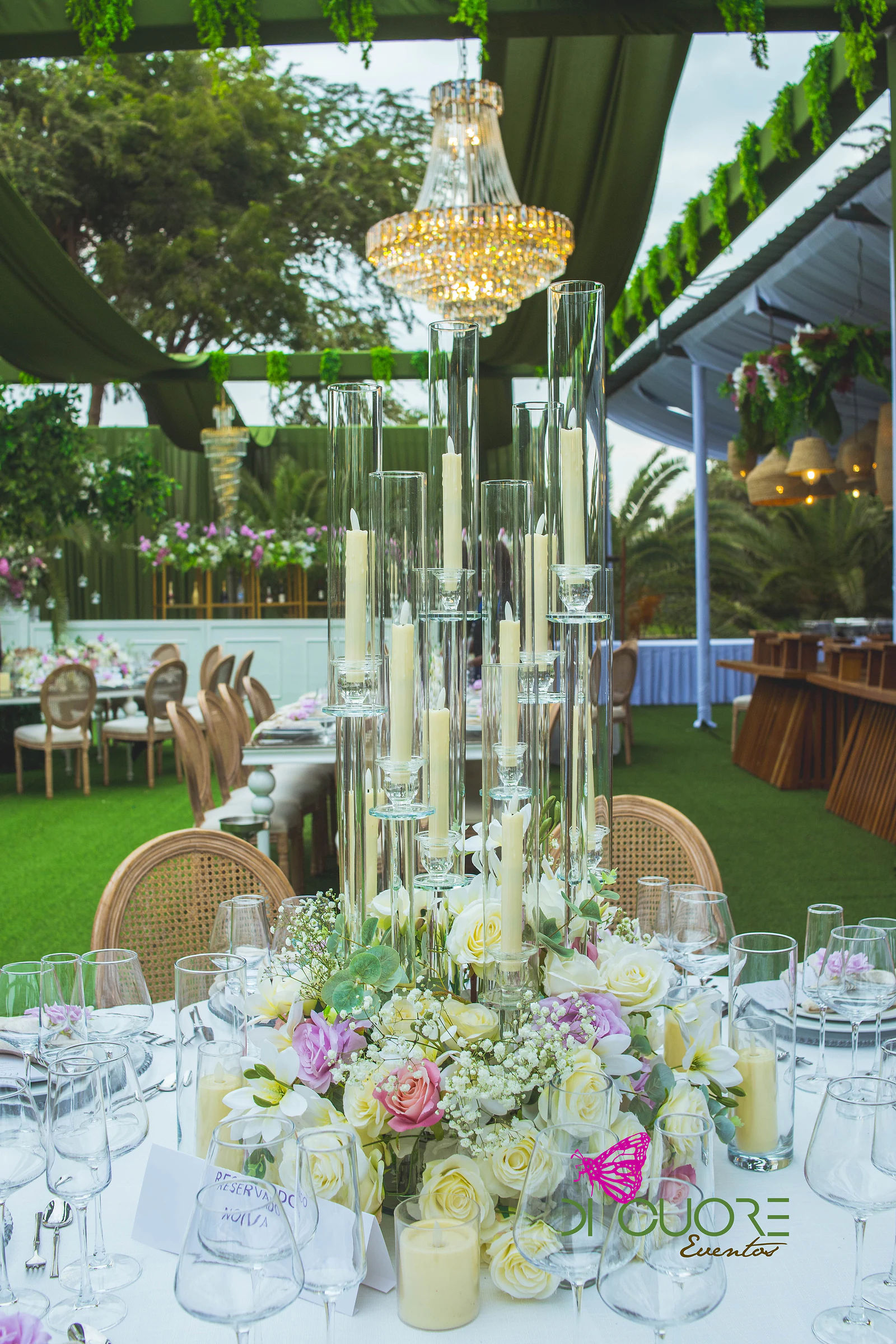 Centro de mesa com candelabros altos de velas em cilindros de vidro, rosas e flores brancas, e um lustre ao fundo