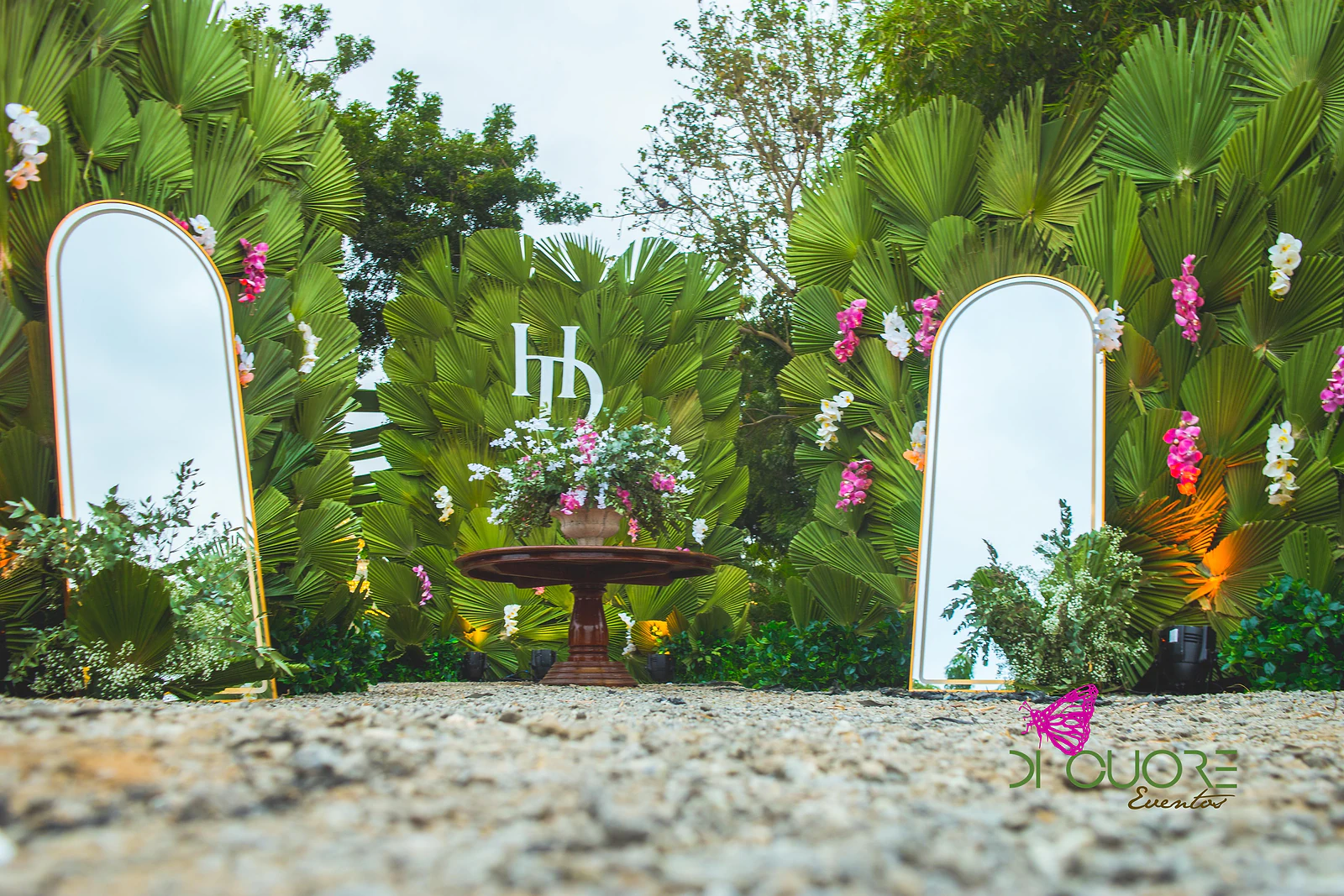 Parede de folhas de palmeira com o monograma HD ao centro, dois espelhos arqueados de moldura dourada e uma mesa redonda de madeira com arranjo de flores, num jardim