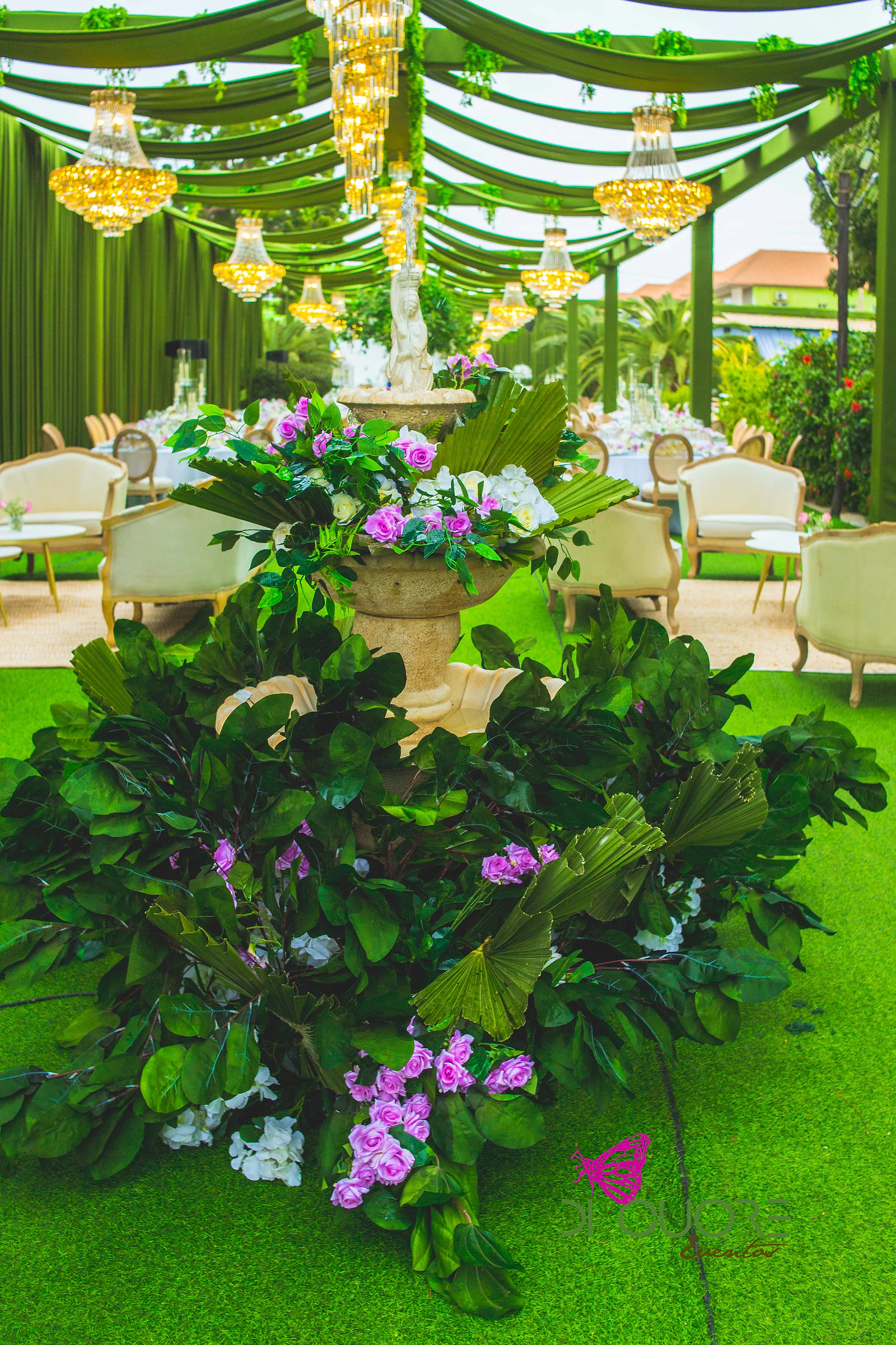 Fonte de pedra coberta de folhas, rosas e flores lilás sobre relva, com sofás cor de creme e lustres dourados ao fundo