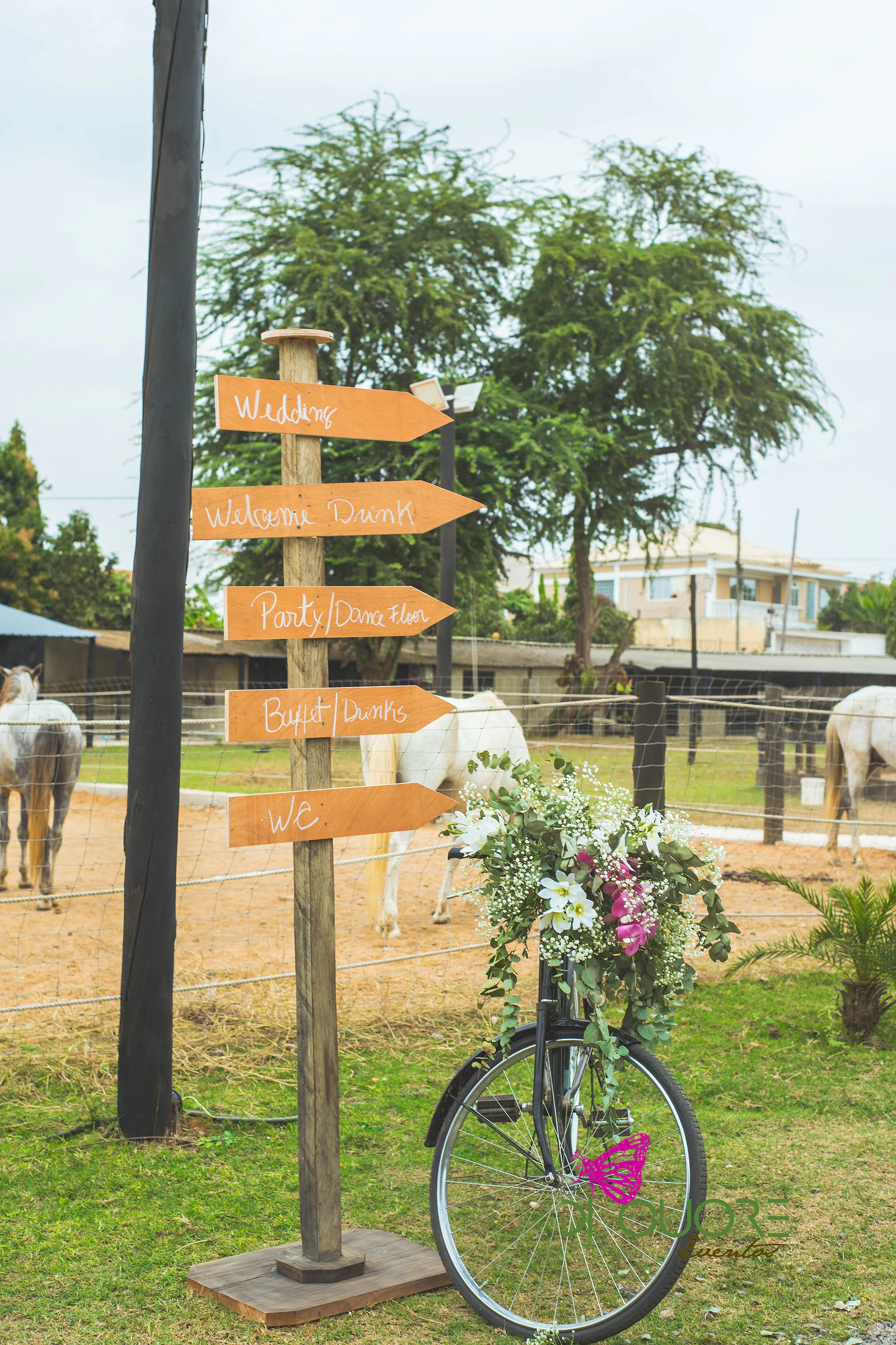 Poste de madeira com placas escritas à mão e uma bicicleta preta com um cesto de flores brancas e cor-de-rosa