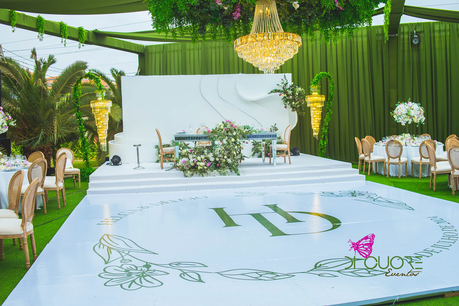 Pista de dança branca com o monograma H&D a verde, degraus e palco branco com mesa de espelho e flores, sob um aro de folhagem e lustre dourado