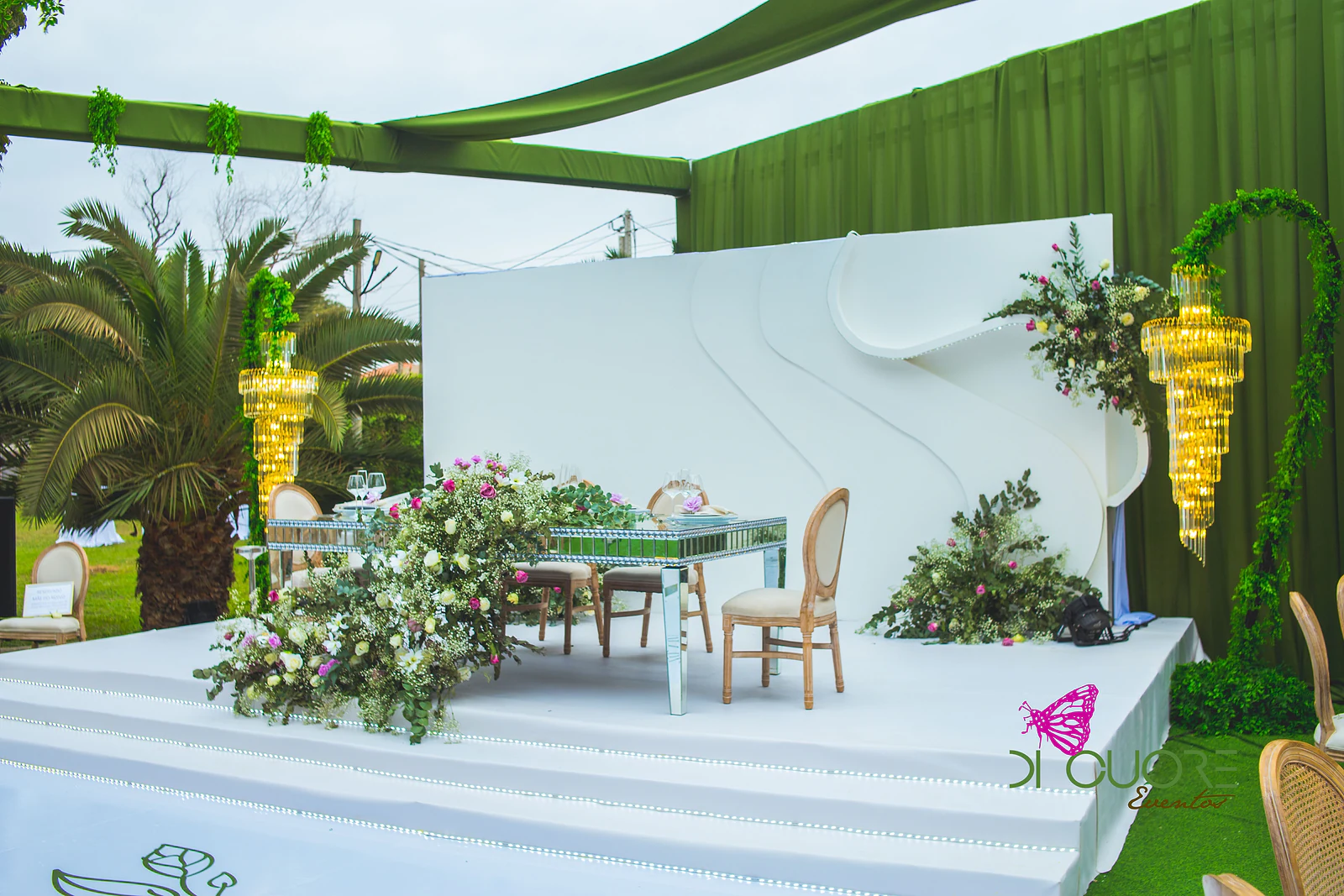 Mesa dos noivos em espelho sobre o palco branco, com uma cascata de flores e folhagem e um lustre dourado ao lado