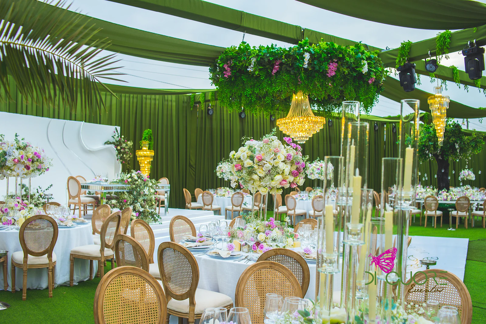 Mesas redondas de toalha branca com cadeiras de palhinha, candelabros altos e ramos de rosas, sob lustres dourados e um aro de folhagem