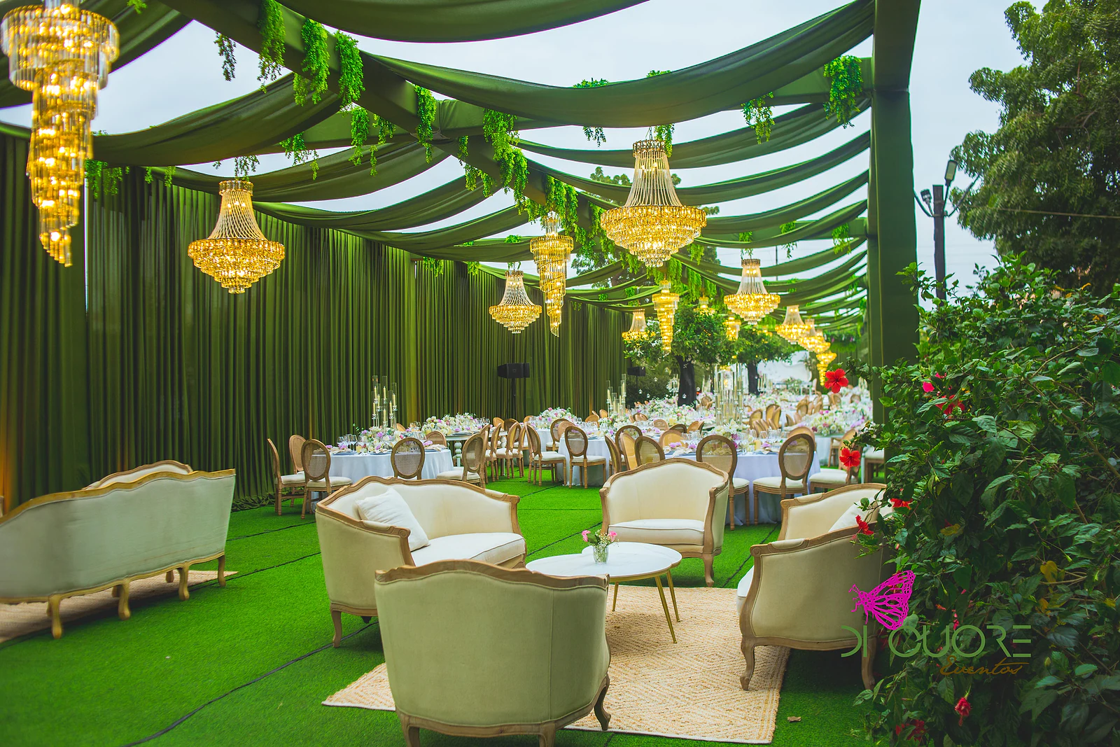 Zona lounge com sofás e cadeirões cor de creme sobre um tapete de fibra, sob panos verdes e lustres dourados
