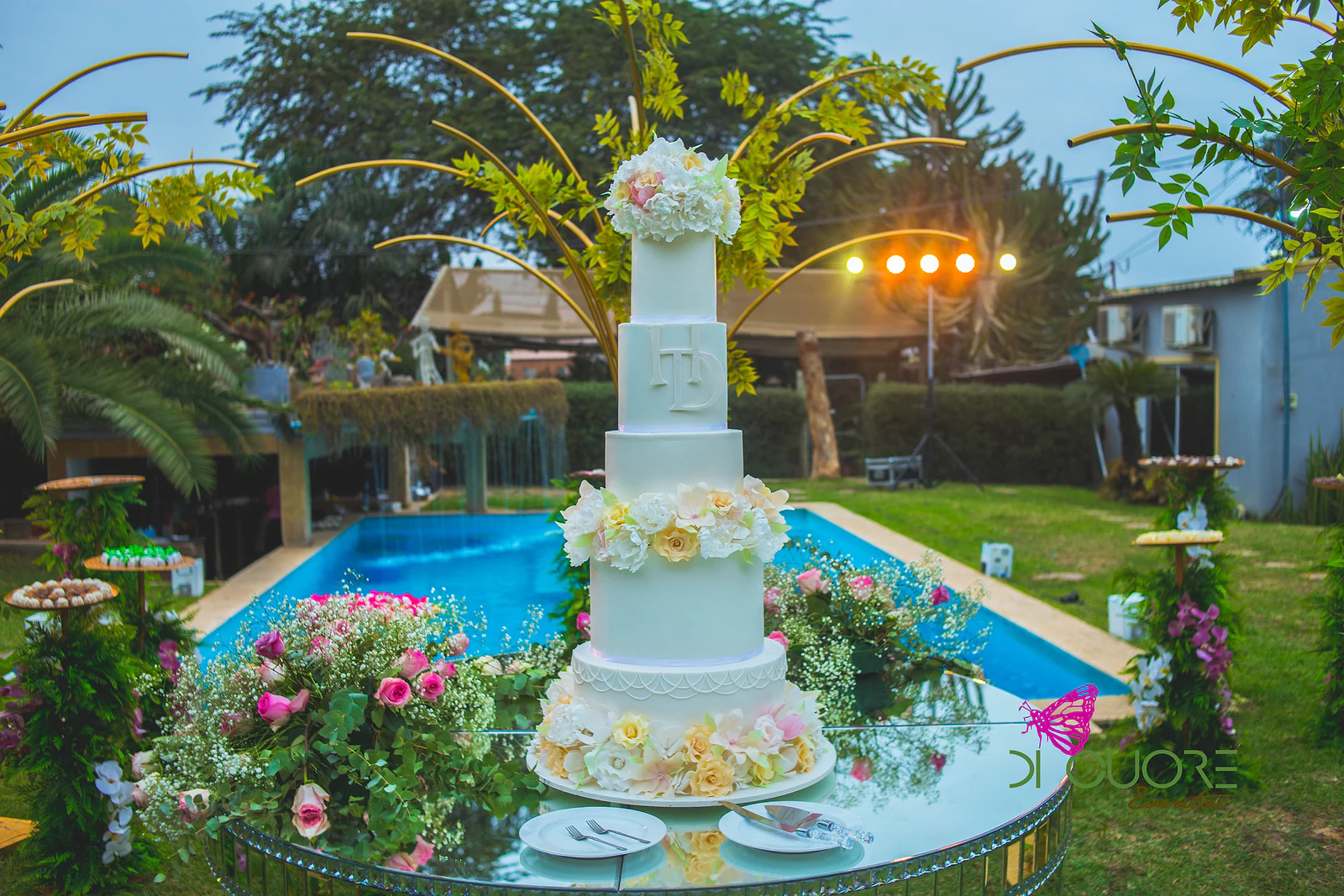 Bolo branco de vários andares sobre mesa de espelho com rosas, junto à piscina e entre palmeiras douradas