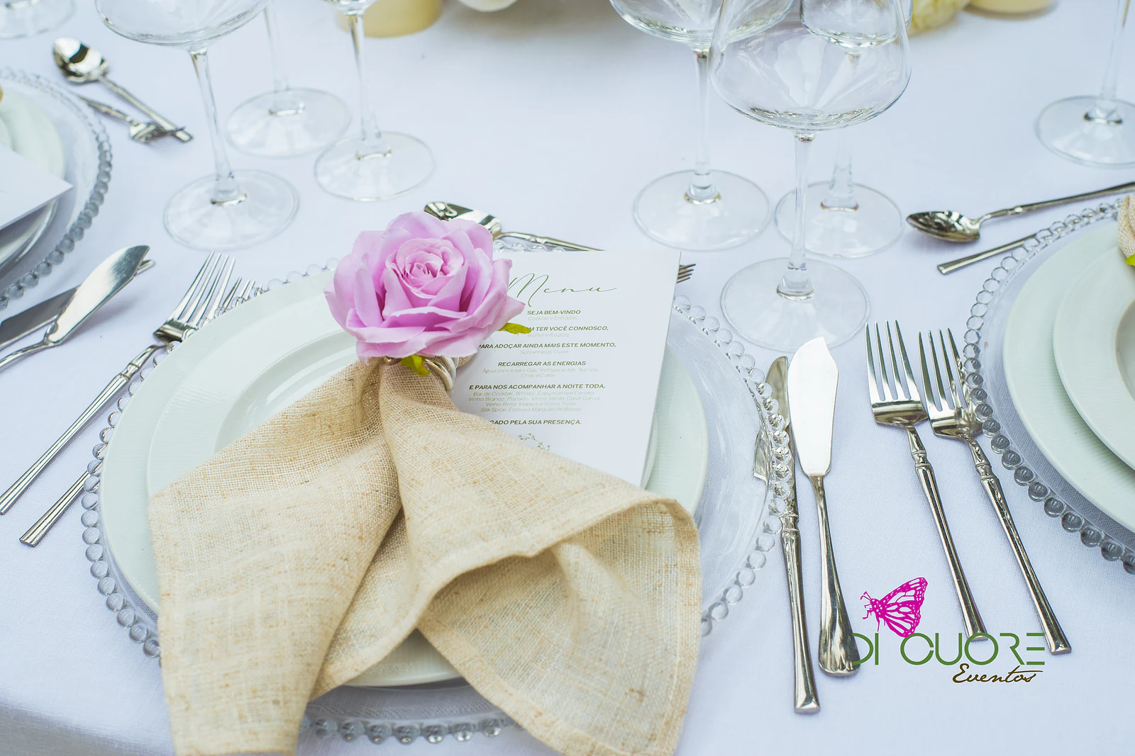 Lugar à mesa com guardanapo de serapilheira, rosa lilás, ementa e prato de cristal sobre toalha branca