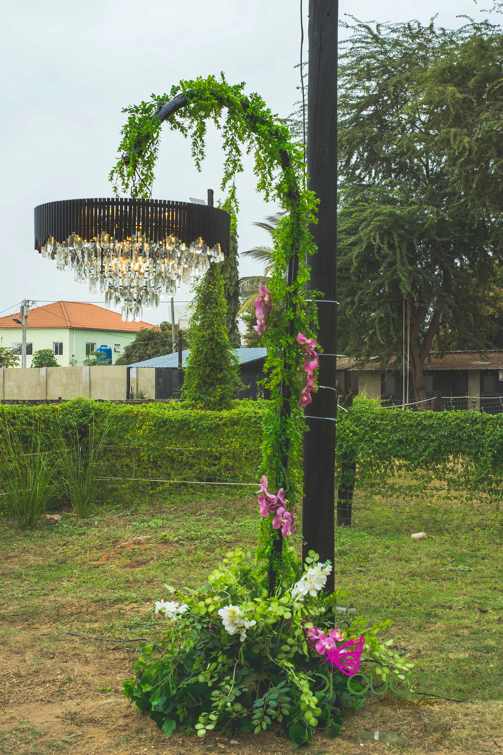 Arco de folhagem com orquídeas rosa e um lustre negro de cristal suspenso, com uma coluna de flores e verde na base