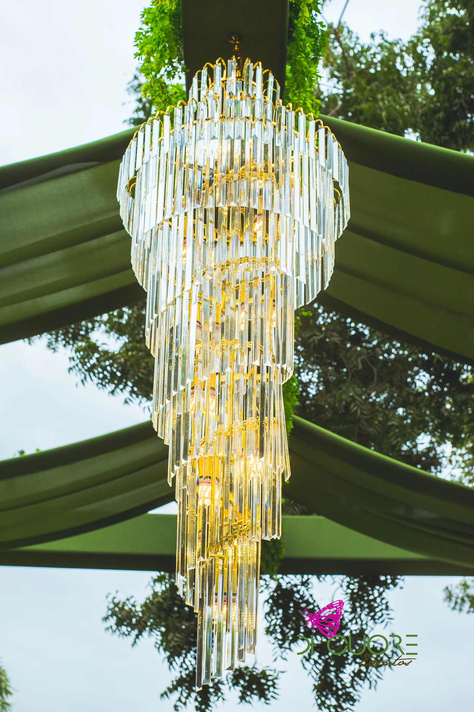 Lustre de cristal dourado em cascata, visto de baixo, entre panos verde-oliva e folhagem