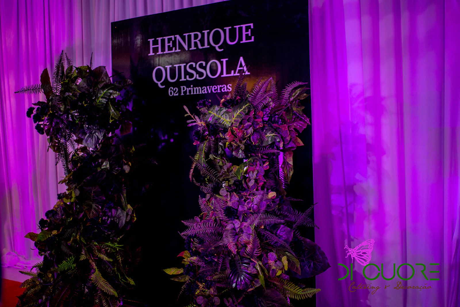 Painel preto com “Henrique Quissola, 62 Primaveras” em letras claras, ladeado por folhagem e cortinados brancos