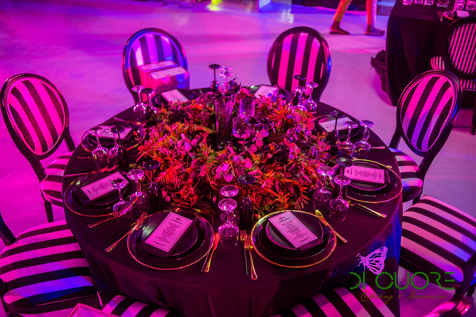 Mesa redonda de toalha preta com centro de folhagem e flores vermelhas, pratos pretos com menu e cadeiras de riscas pretas e brancas