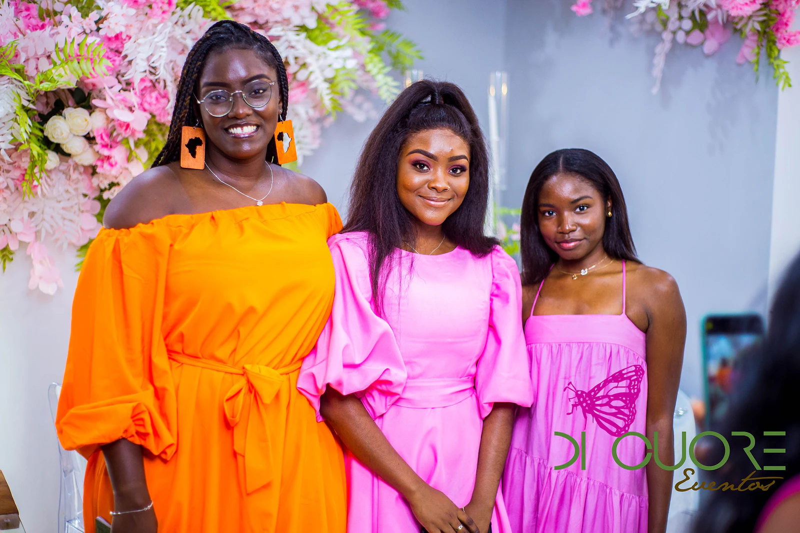 Três convidadas, de vestido laranja e cor-de-rosa, a sorrir diante de flores cor-de-rosa e brancas