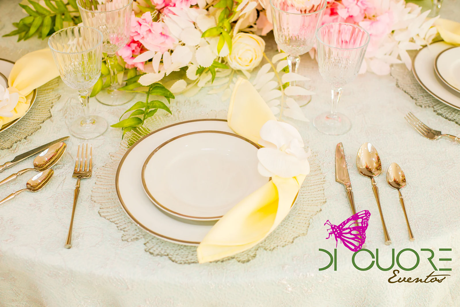 Prato branco com friso dourado sobre toalha verde-água, com guardanapo amarelo-pálido, talheres dourados e flores cor-de-rosa