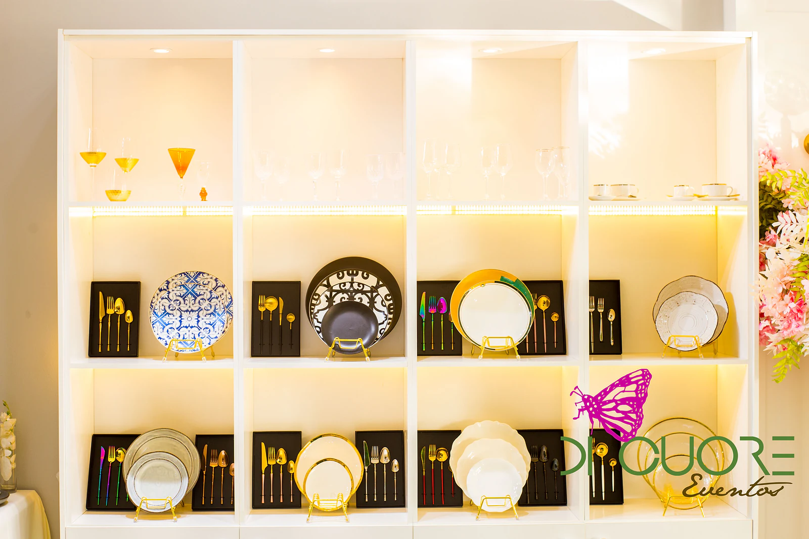 Estante branca iluminada com pratos decorativos, talheres dourados e pretos e taças coloridas