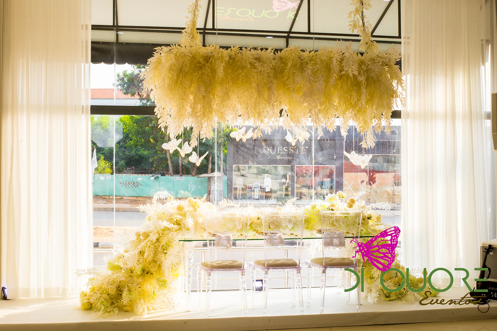 Instalação suspensa em tons de creme sobre uma mesa de vidro com cadeiras transparentes e flores brancas a cair para o chão, junto à janela