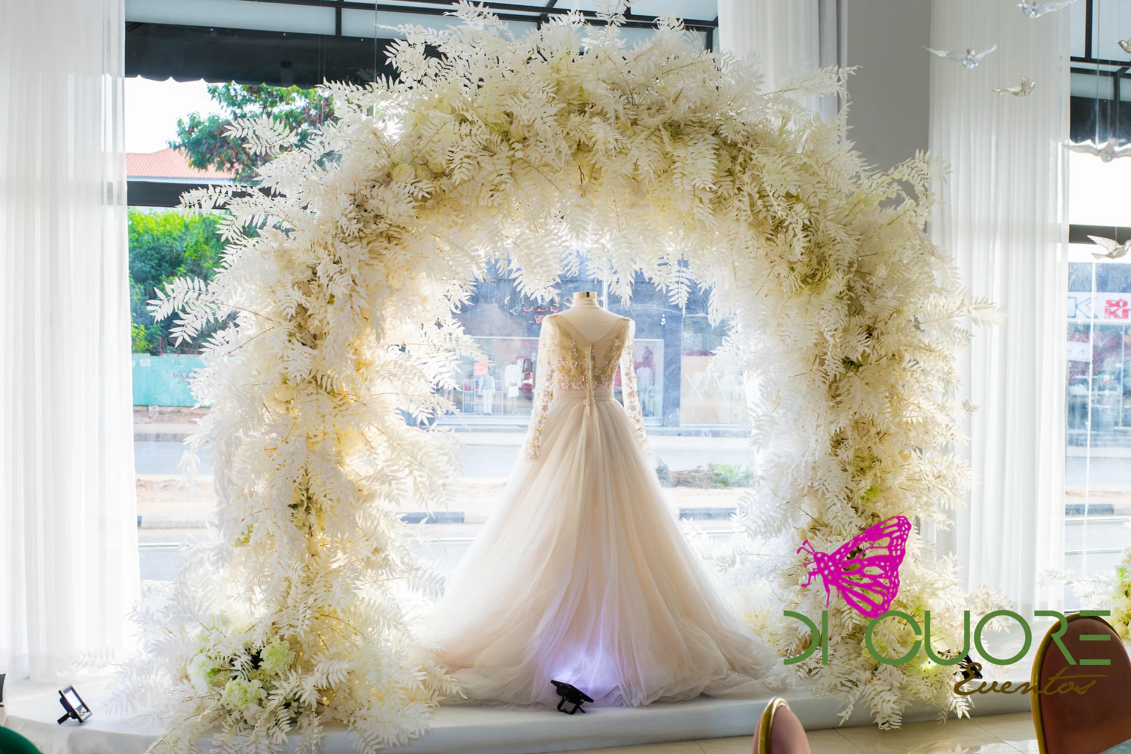 Arco redondo de folhagem e flores brancas a emoldurar um vestido de noiva num manequim, em frente à janela