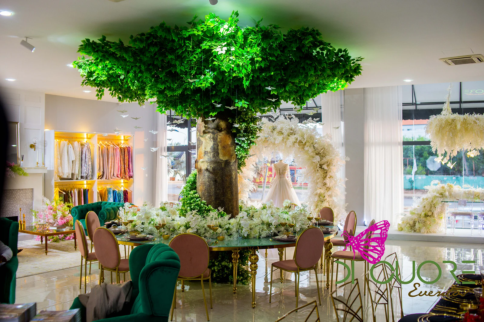 Árvore de folhagem verde sobre uma mesa redonda posta com flores brancas, entre cadeiras cor-de-rosa e cadeirões de veludo verde
